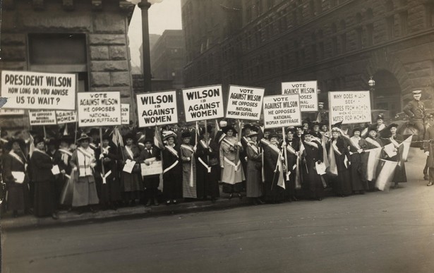 Women suffrage activists wearing suffrage sashes demonstrating with signs at city street corner in 1916. (Wikimedia)