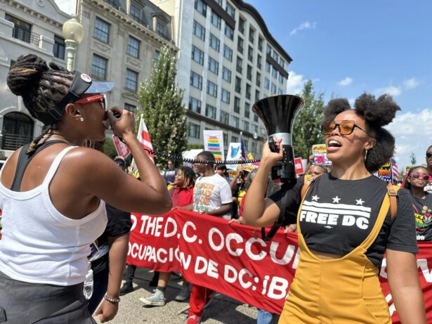 A woman with a megaphone and a FREE DC shirt marches in front of a banner that partially reads "the DC Occupation"